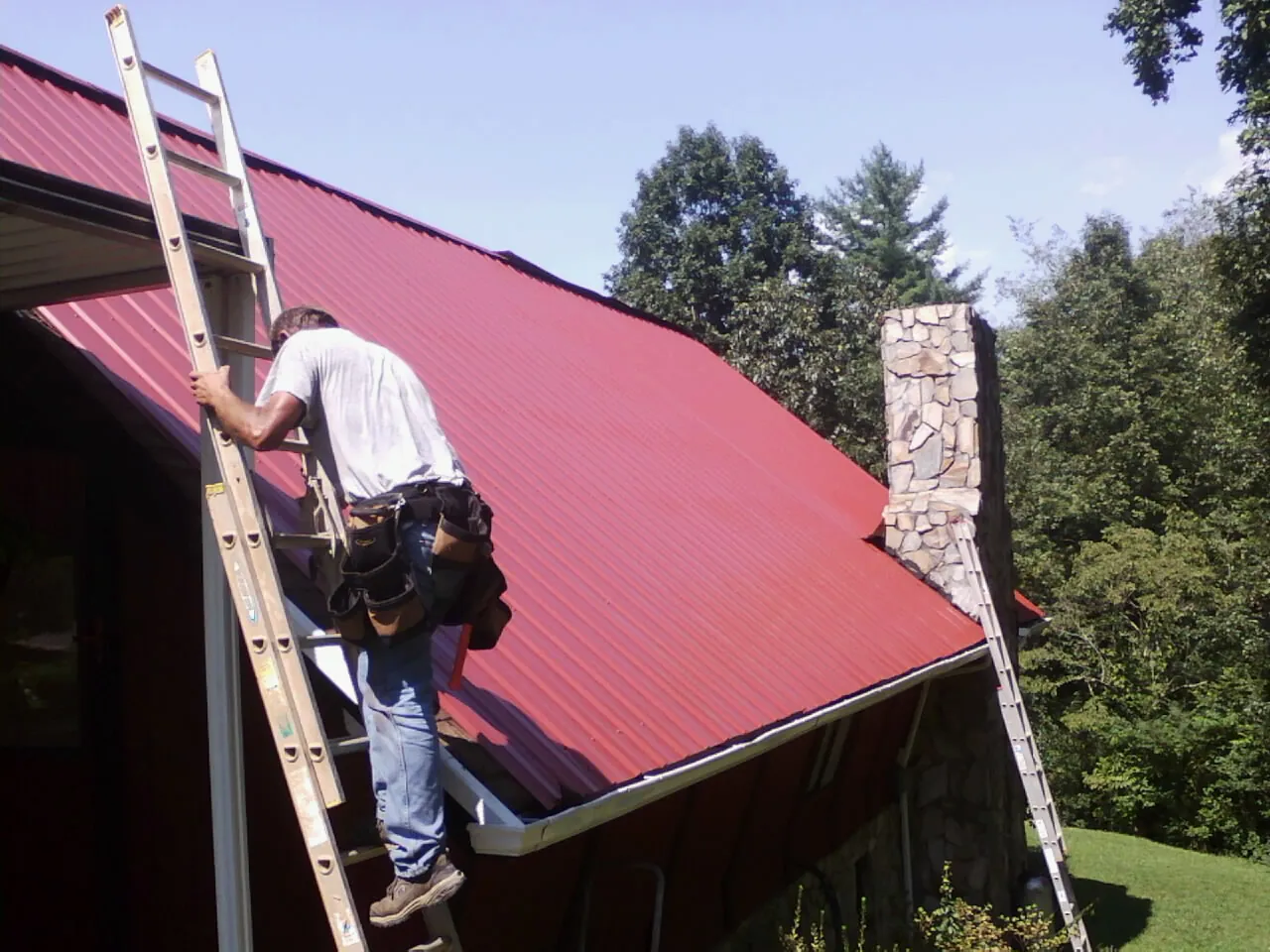 Roofer on a ladder beside a red metal roof and stone chimney — Soffit & Fascia Repair by Roofing Brothers in Fillmore